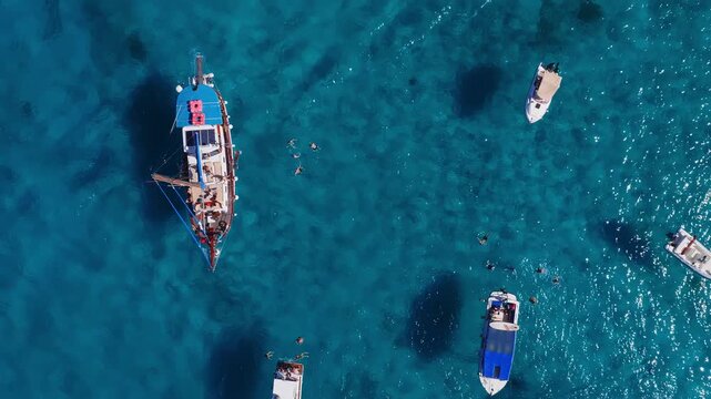 Aerial top down view at Blue Lagoon, Comino, Malta. Yachts, wooden boats, motorboats, and swimmers move over clear water with seagrass patches and bright highlights.