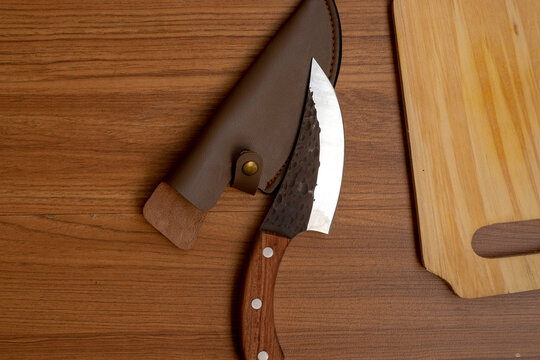 A top-down view shows a hand-forged knife with a hammered blade finish and wooden handle, partially inside its brown leather sheath on a wood table beside a cutting board.