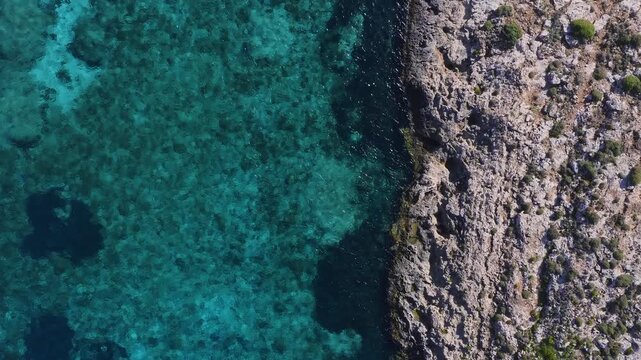 Aerial top down view of Comino Island, Malta, with rugged limestone coast right and clear turquoise sea left. Underwater rocks and dark pools show color gradations.