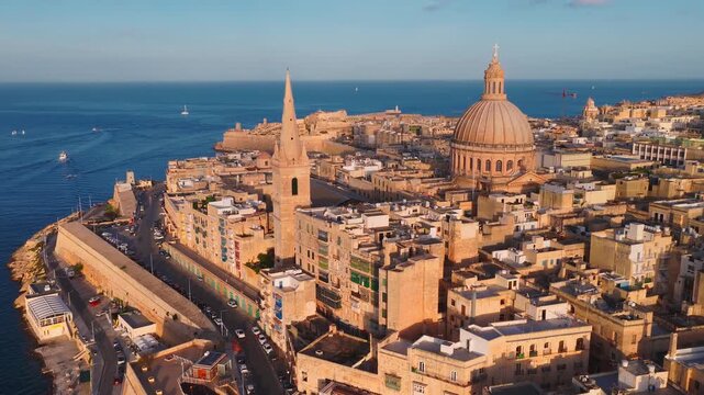 Aerial view of Valletta, Malta at dusk shows basilica dome, pro cathedral spire, bastioned walls, seawalls, boats, narrow streets, balconies, terraces, and coast roads.
