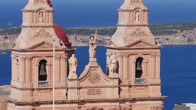 High aerial shot pans across Mellieha Parish Church facade in northern Malta, twin bell towers, red dome, Latin text, boats and yacht on cobalt sea in bright daylight