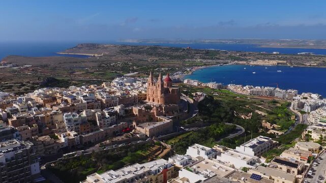 Sunlit aerial view of Mellieha, Malta centers on the sandstone parish church, red dome and twin spires, with Ghadira Bay, boats, moving cars, and Comino and Gozo visible.
