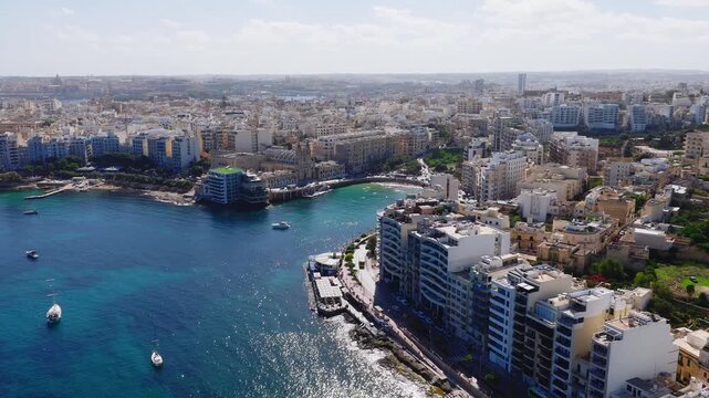 Aerial view shows Sliema waterfront in Malta with limestone blocks, hotels, rocky promenade, crescent bay, floating lido, sailboats, midday light, people and traffic