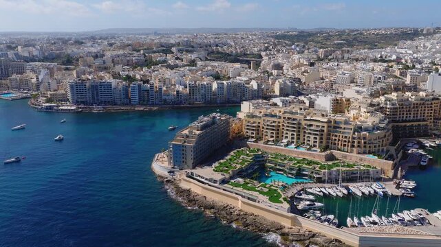 Aerial view of Sliema, Malta shows limestone waterfront apartments, hotels, marina, promenade, turquoise water, yachts, boats, rooftop pools, and gentle waves.