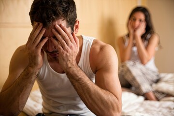Upset man with relationship problems in bedroom with concerned woman behind