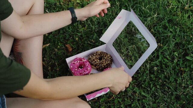 Woman sitting on the grass opens a pink box with chocolate and strawberry donuts, enjoying the sweet aroma on a summer day. Relaxed outdoor snack moment. 4k horizontal footage
