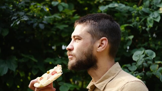 Close-up of a bearded man enjoying a slice of pizza with cheese, ham, tomato, and olives in the park on a sunny day. Relaxed summer vibe. 4k horizontal footage