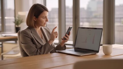A focused woman in a business setting examines her smartphone with a laptop open Stock Video - Powered by Adobe