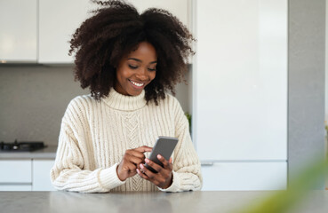 Smiling black woman texts on smartphone from her modern kitchen counter. She wears a cozy sweater and uses a mobile device for communication or shopping.