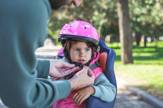 Father fastening pink helmet on his little daughter sitting in a child bicycle seat, both outdoors in a sunny park, concept of protection, safety, parental care and outdoor family activity
