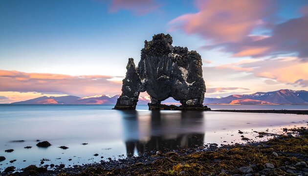 A majestic dark rock formation, its reflection shimmering in calm water, against a dramatic sunset sky. Mountains and coast in the background
