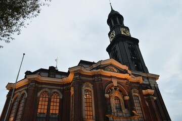 The majestic baroque facade of St. Michael's Church (Hauptkirche Sankt Michaelis) in Hamburg, Germany, illuminated with warm golden lights against a moody, overcast twilight sky.