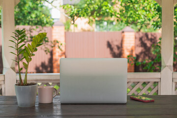 Modern workspace outdoors with laptop on wooden table, potted green plant, coffee mug and smartphone, surrounded by garden greenery, concept of remote work, digital lifestyle and productivity