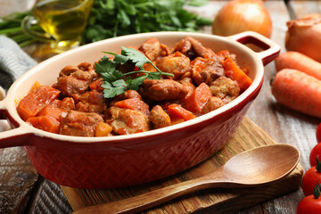 Delicious cooked stew in baking dish, fresh ingredients and spoon on wooden table, closeup