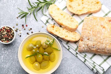 Fresh cut ciabatta, oil, olives, rosemary and peppercorns on grey textured table, flat lay