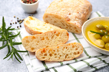 Fresh cut ciabatta, oil, olives, rosemary and peppercorns on grey textured table, closeup