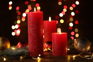 Burning candles, fir tree branches, baubles and festive lights on wooden table against black background with blurred lights, closeup. Bokeh effect