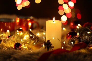 Burning candle, fir tree branches, baubles, cones and festive lights on faux fur against black background with blurred lights, closeup. Bokeh effect