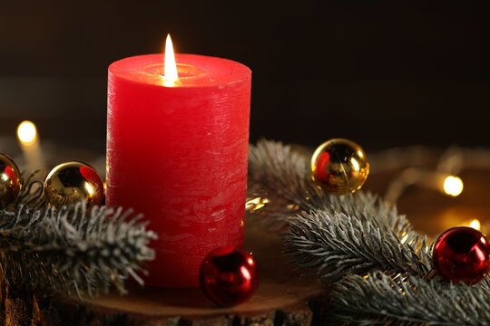 Burning candle, fir tree branches and baubles on table against black background, closeup. Christmas greeting card