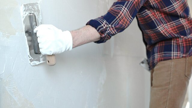 Unknown male construction worker wearing protective gloves smoothing plaster on interior wall during home renovation, applying material with professional trowel technique, close-up