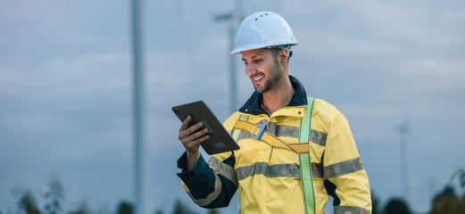 Male engineer in smiling while using digital tablet at wind farm during evening, monitoring renewable energy system, man worker at wind turbine. © eakgrungenerd