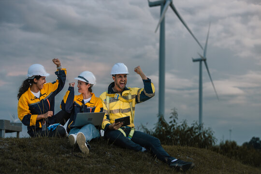 Happy team of engineers celebrating project success at wind farm, using digital devices for data analysis, teamwork and renewable energy planning in outdoor environment.