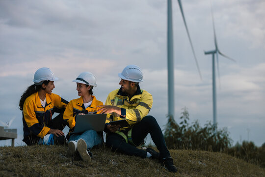 Group of engineers wearing safety helmets and uniforms working together outdoors at wind farm, using laptop and tablet for data analysis, teamwork and sustainable energy project planning.