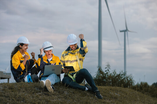 Happy team of engineers celebrating project success at wind farm, using digital devices for data analysis, teamwork and renewable energy planning in outdoor environment.