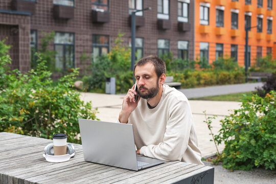 Bearded man sitting outdoors with laptop, talking on smartphone, coffee and headphones nearby, concept of remote work, communication, freelance and modern digital lifestyle