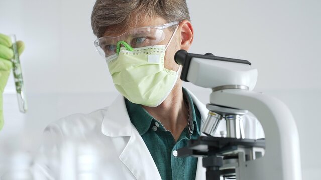 Adult male scientist wearing green mask, gloves and goggles using microscope and analyzing sample in test tube in a laboratory, developing new drugs and vaccines. Medicine and science concept