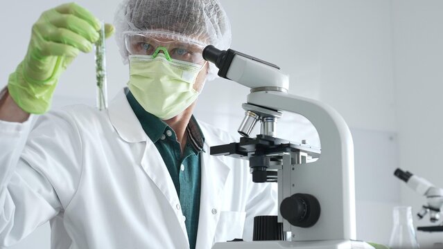 Adult male scientist wearing green protective gear looking at plant samples in test tubes and using a microscope in modern laboratory for botanical research. Medicine, healthcare and science concept