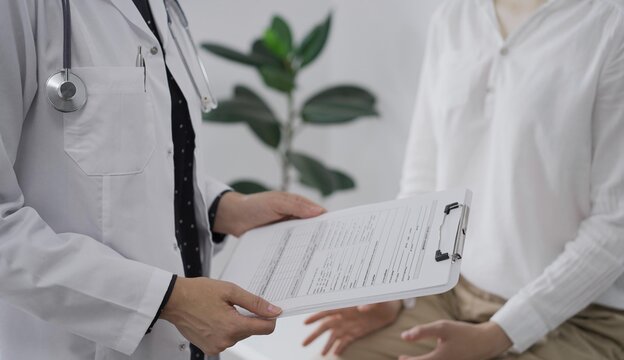 Doctor and a patient discussing something. The female physician, wearing a white medical coat is keeping clipboard with a medical notes besides a young woman during a consultation in the clinic