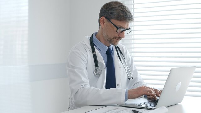 A male doctor in glasses and a lab coat with a stethoscope is typing on a laptop keyboard while sitting at a table in a bright medical office