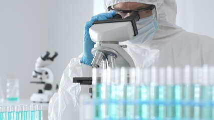Medical scientist wearing protective suit using microscope while analyzing samples, conducting research in laboratory environment. Close up of test tubes with blue liquid - Powered by Adobe