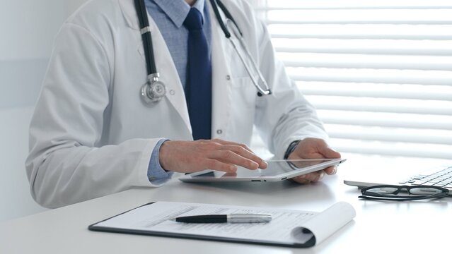 Doctor wearing lab coat and stethoscope using digital tablet while sitting at desk in medical office with laptop, eyeglasses, and medical chart nearby. Medicine and health care concept