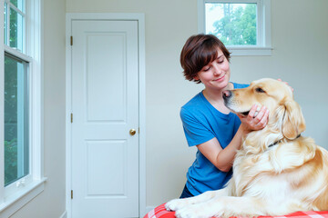 Caucasian young adult woman gently petting Golden Retriever dog indoors, smiling and making eye contact with animal, natural light coming through windows, relaxed interaction