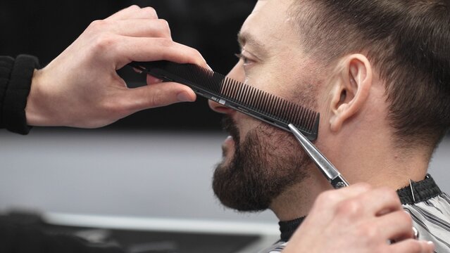 Skilled barber carefully trimming client's beard with precision scissors and sharp comb, working inside contemporary grooming salon
