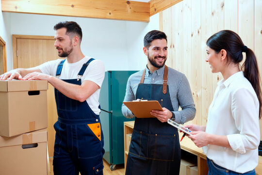 Three young adult workers, two Caucasian men and one Asian woman, collaborating in warehouse, one man stacking cardboard boxes while others discussing logistics using clipboard and tablet