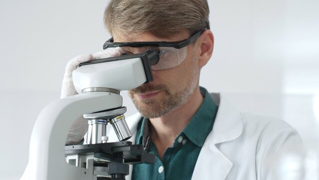 Senior man scientist wearing protective glasses and lab coat analyzing samples with a microscope in laboratory setting. Medicine, health care and science concept