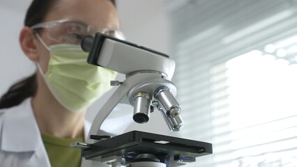 Female scientist wearing green mask and protective glasses using microscope in laboratory, doing scientific research and looking for a vaccine. Medicine, healthcare and science concept
