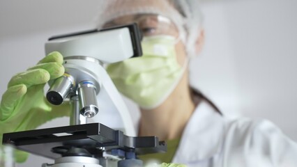 Female scientist wearing green mask and protective glasses using microscope in laboratory, doing scientific research. Medicine, healthcare and science concept