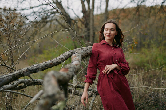 A young woman in a red dress leans against a fallen tree on a grassy hillside, captured with authentic, genuine expression and realistic textures that evoke quiet natural confidence.