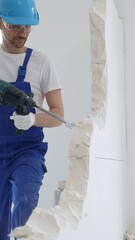 Male construction worker wearing blue overalls, protective helmet and white gloves, is demolishing white wall with rotary hammer drill, generating dust, close up horizontal view
