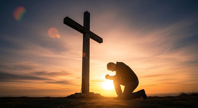 Silhouette of a man praying at the foot of the cross at sunset.