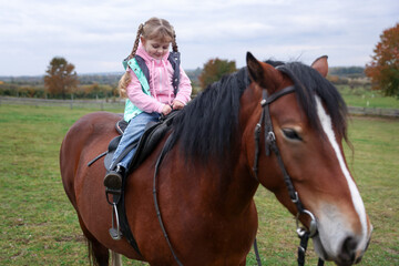 Fototapeta premium Equine assisted therapy. Little girl riding beautiful horse outdoors. Lovely domesticated pet
