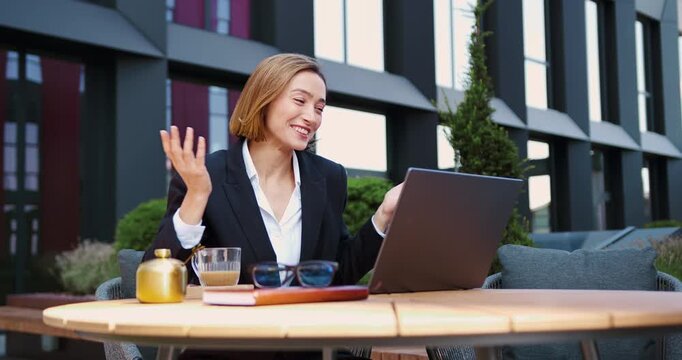 Handsome woman using laptop having an online meeting with client or business partner during lunch break of the cafe. Smiling mature successful businesswoman communicates remotely on a video call near