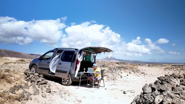 Mature digital nomad sitting at a table with a laptop, enjoying a freelance lifestyle working remotely from a volcanic beach with his customized minivan parked nearby