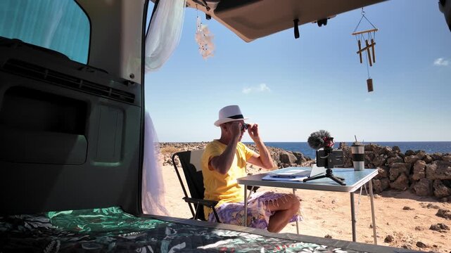 Mature man working as a digital nomad, enjoying the freedom of freelancing from his minivan office with a beautiful volcanic sea view while taking a break to sunbathe and relax