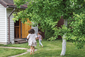 Obraz premium Two barefoot children running across a green garden toward a small wooden house on a sunny day, surrounded by lush trees, representing childhood, freedom, and outdoor playtime