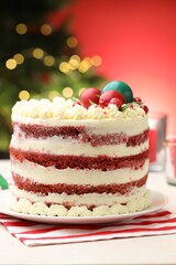 Delicious sponge cake with Christmas decor on white table against red background with blurred lights, closeup. Bokeh effect